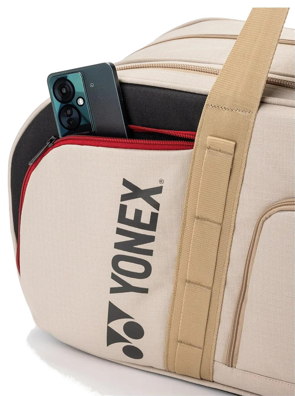 A beige Yonex BA72631WEX Gearlogic Tournament Badminton Bag with a tan strap and red zipper pocket is shown partially unzipped, revealing a green smartphone inside.