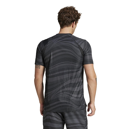 A curly-haired person stands with their back to the camera, showcasing an ADIDAS Men's Club Graphic Badminton T-Shirt in black, made from 100% recycled materials. The athletic outfit features AEROREADY technology and includes both a patterned short-sleeved shirt and matching shorts.