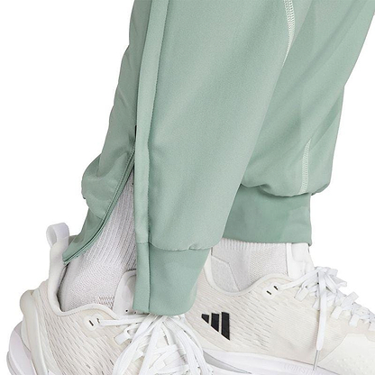 A person wearing adidas Melbourne Men's Pro Badminton Pants in green, equipped with AEROREADY technology, paired with white sneakers.