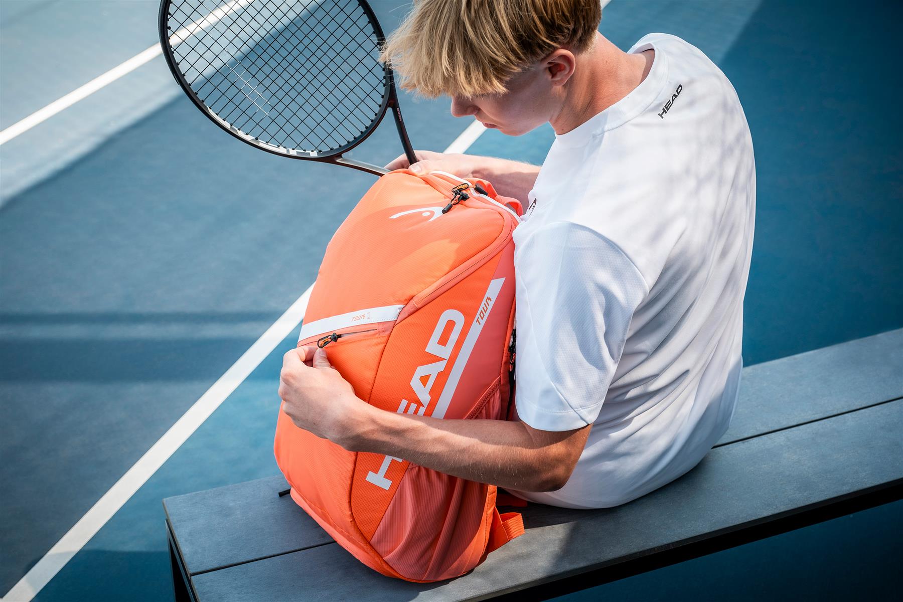 A tennis player in a white shirt sits on a blue court, holding a HEAD Tour 25L Badminton Backpack - Orange and a racket, opening or closing the backpack’s zipper.
