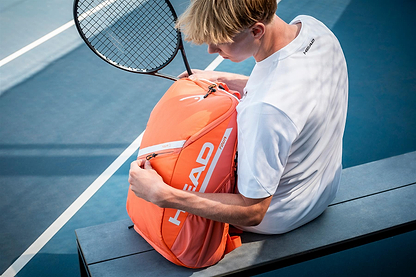 A tennis player in a white shirt sits on a blue court, holding a HEAD Tour 25L Badminton Backpack - Orange and a racket, opening or closing the backpack’s zipper.