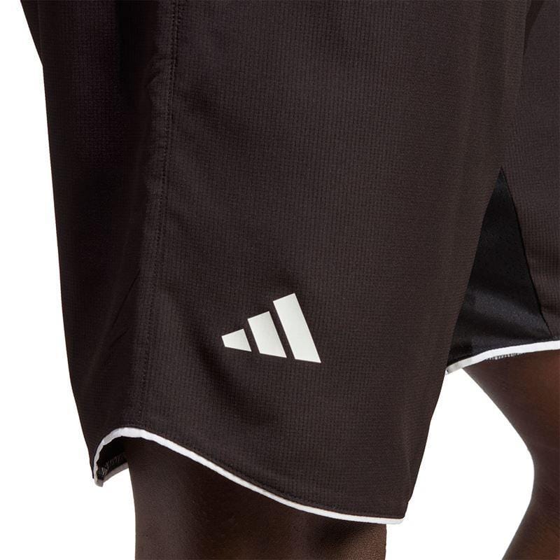 Close-up image of a person wearing the ADIDAS Mens Club 7" Badminton Shorts - Black, featuring a white geometric adidas logo on the side. These shorts incorporate AEROREADY technology and are accented with a white trim along the hem.