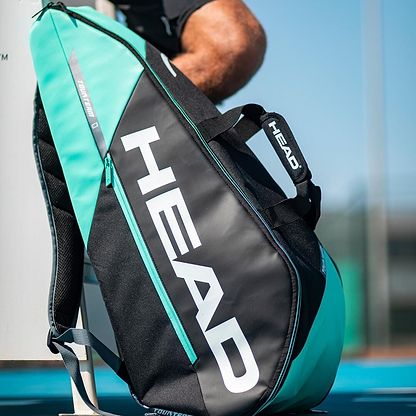 A black and mint HEAD Tour Team 6R Combi racket bag, featuring a prominent white logo and equipped with HEAD's CCT+ climate control technology, is leaning against a bench on the tennis court. Nearby, a person is partially visible, resting after an intense match.
