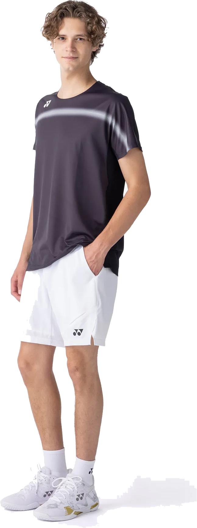 A young man stands with one hand in his pocket, wearing a Yonex 10711YX Crew Neck Mens Badminton T-Shirt in Black, along with white shorts, socks, and athletic shoes—all branded with Yonex logos—against a white background.