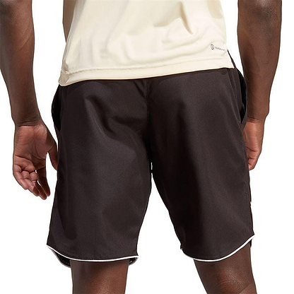 A person wearing a light-colored AEROREADY athletic shirt and the ADIDAS Men's Club 7" Badminton Shorts in black with white trim stands with their back to the camera, hands relaxed by their sides. The plain white background accentuates the sleek design of their attire.