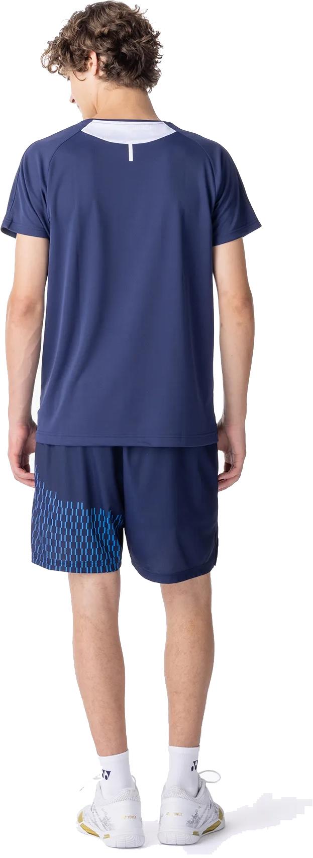 A young man faces away from the camera, wearing a Yonex 10713EX Crew Neck Men's Badminton T-Shirt in dark navy, paired with patterned navy shorts, white socks, and white athletic shoes designed for superior moisture management.