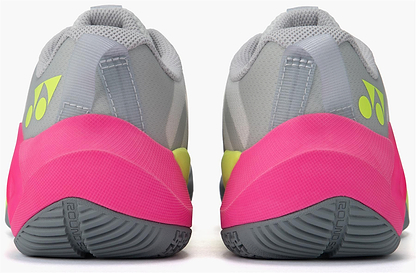 The Yonex Subaxia GT Women's Badminton Shoes in light grey feature bright pink heel accents, yellow Yonex logos, textured dark grey soles, and advanced GRPHT THRTTL technology for enhanced performance.