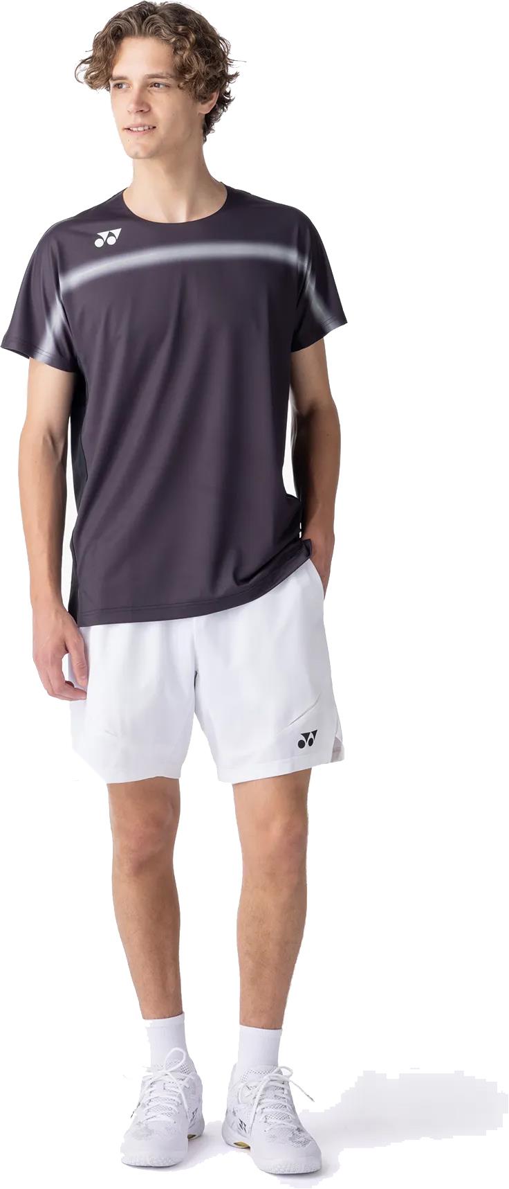 A young man in a Yonex 10711YX Crew Neck Men's Badminton T-Shirt (Black), paired with white shorts, socks, and sneakers, stands with one hand in his pocket, gazing to the side.