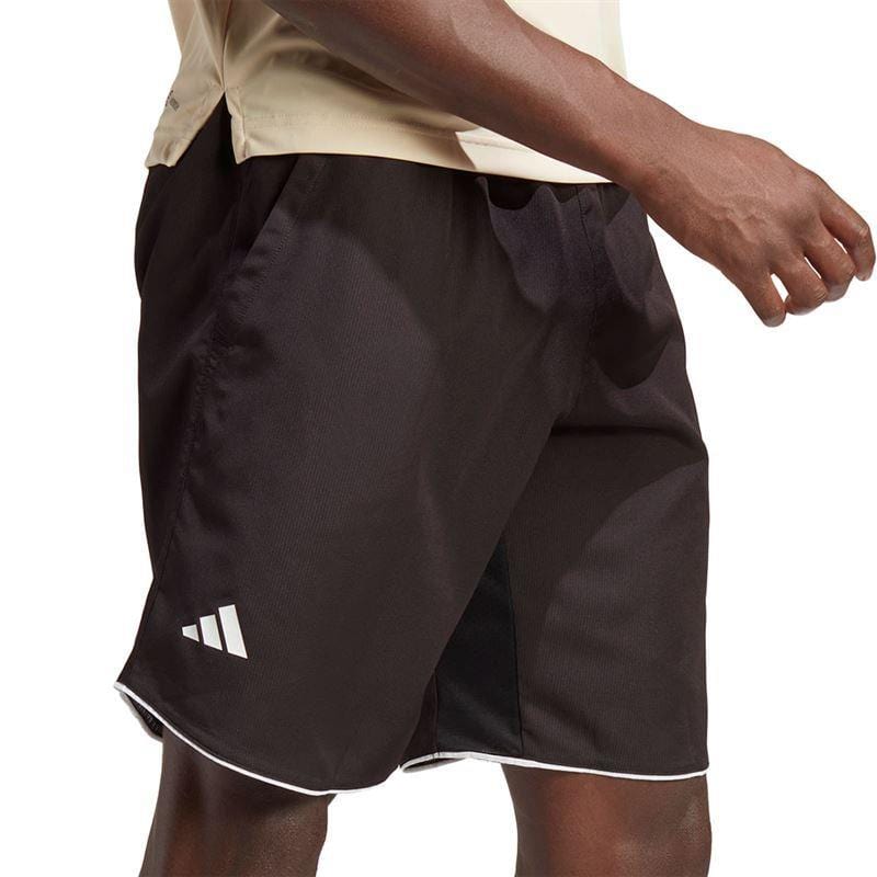 A person in a beige shirt is wearing black adidas Men's Club 7" Badminton Shorts, highlighting the iconic white logo on the side. The shorts, crafted from lightweight ripstop fabric, feature a white trim at the bottom. The image emphasizes the lower half of the body against a plain white background.