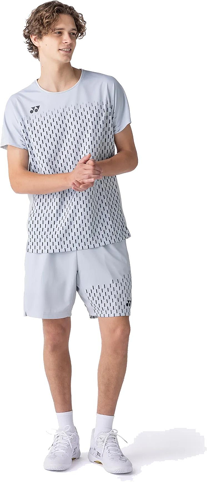 A young man wears a light gray athletic set with black dash patterns, featuring a Yonex 15241EX Knit Mens Badminton Shorts in Silver Grey and white sneakers. He looks to the side, hands clasped.
