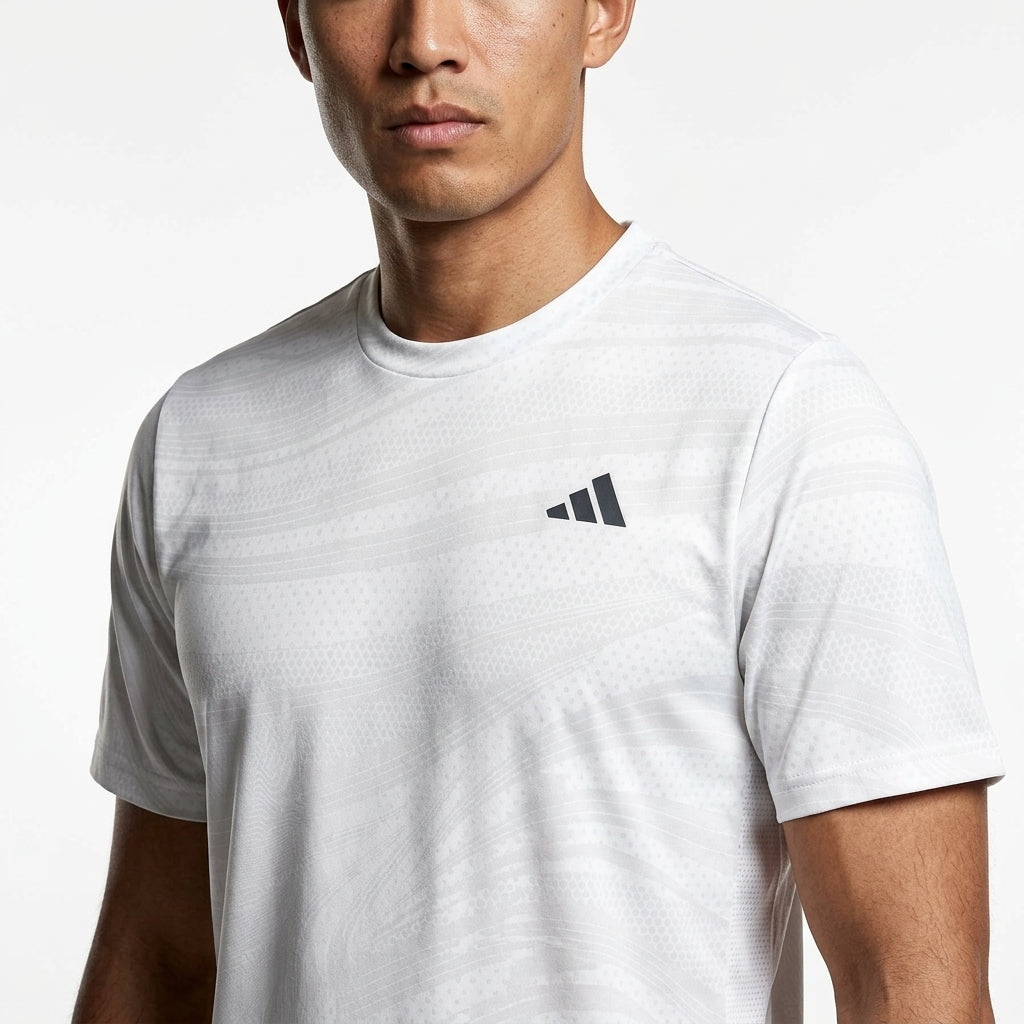 A man wears the adidas Mens Club Graphic Badminton T-Shirt in white/grey, a fitted short-sleeve sportswear top made from recycled materials with moisture-wicking fabric and a small black adidas logo on the chest.