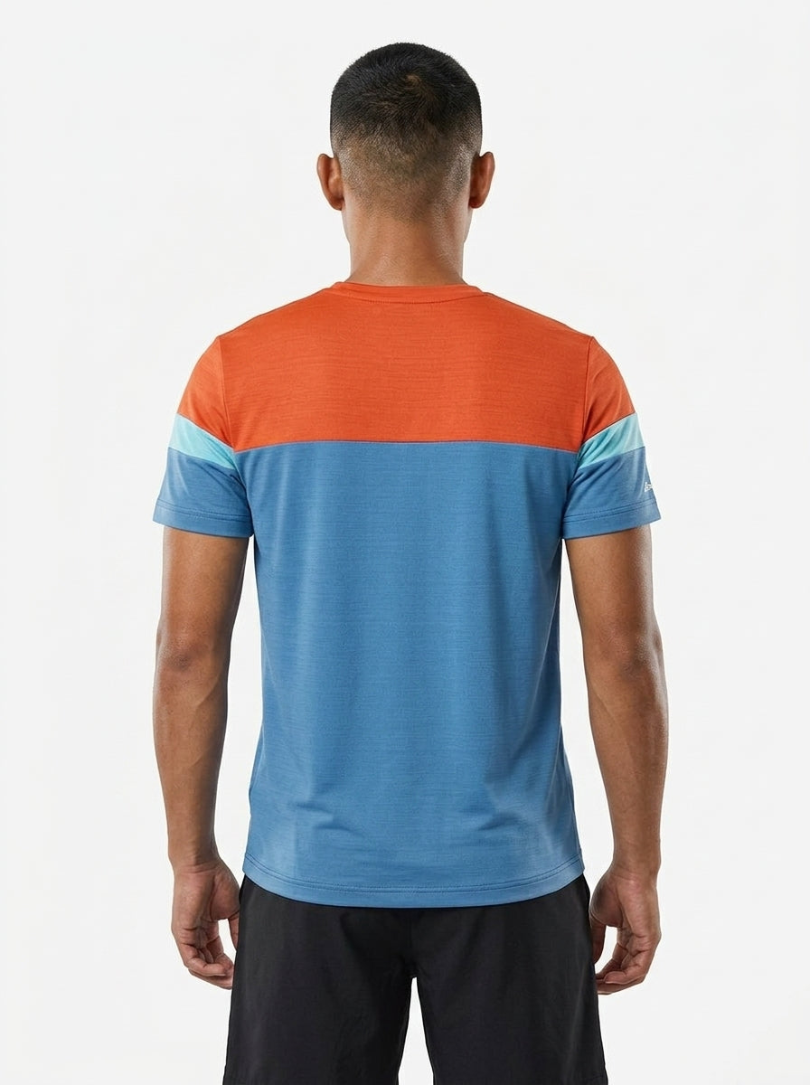 A man faces away from the camera, wearing a Babolat Crew Neck Mens Badminton T-Shirt - Seaport with an orange top, light blue sleeve stripes, blue lower half, and black shorts against a plain white background.