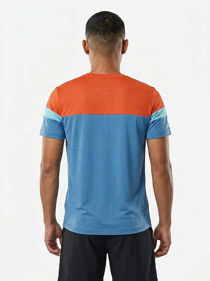 A man faces away from the camera, wearing a Babolat Crew Neck Mens Badminton T-Shirt - Seaport with an orange top, light blue sleeve stripes, blue lower half, and black shorts against a plain white background.