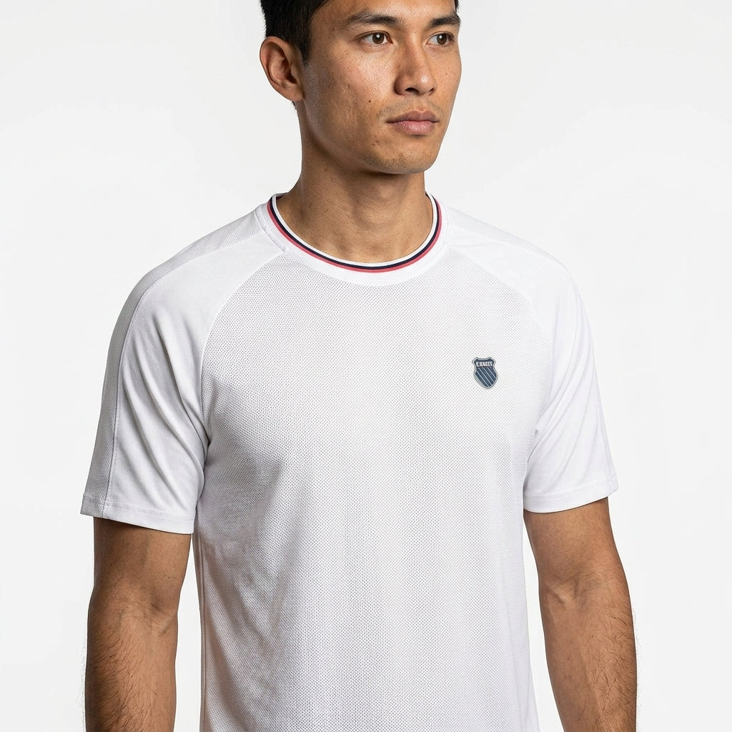 A man wears the K-Swiss Hypercourt Mesh Crew 4 Men's Badminton T-Shirt in white, featuring a small chest logo and a red, white, and blue striped collar, standing against a plain white background.