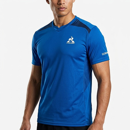A man wears the Le Coq Sportif Pro Mens Badminton T-Shirt in Lapis Blue, featuring short sleeves, black mesh shoulder accents, and branded logos—ideal breathable sportswear from Le Coq Sportif for active days.