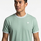 A man wears a sage green Tecnifibre Team Stretch Men's Badminton T-Shirt with white collar and sleeve trim, featuring a small white Tecnifibre logo on the left chest, standing against a plain white background.
