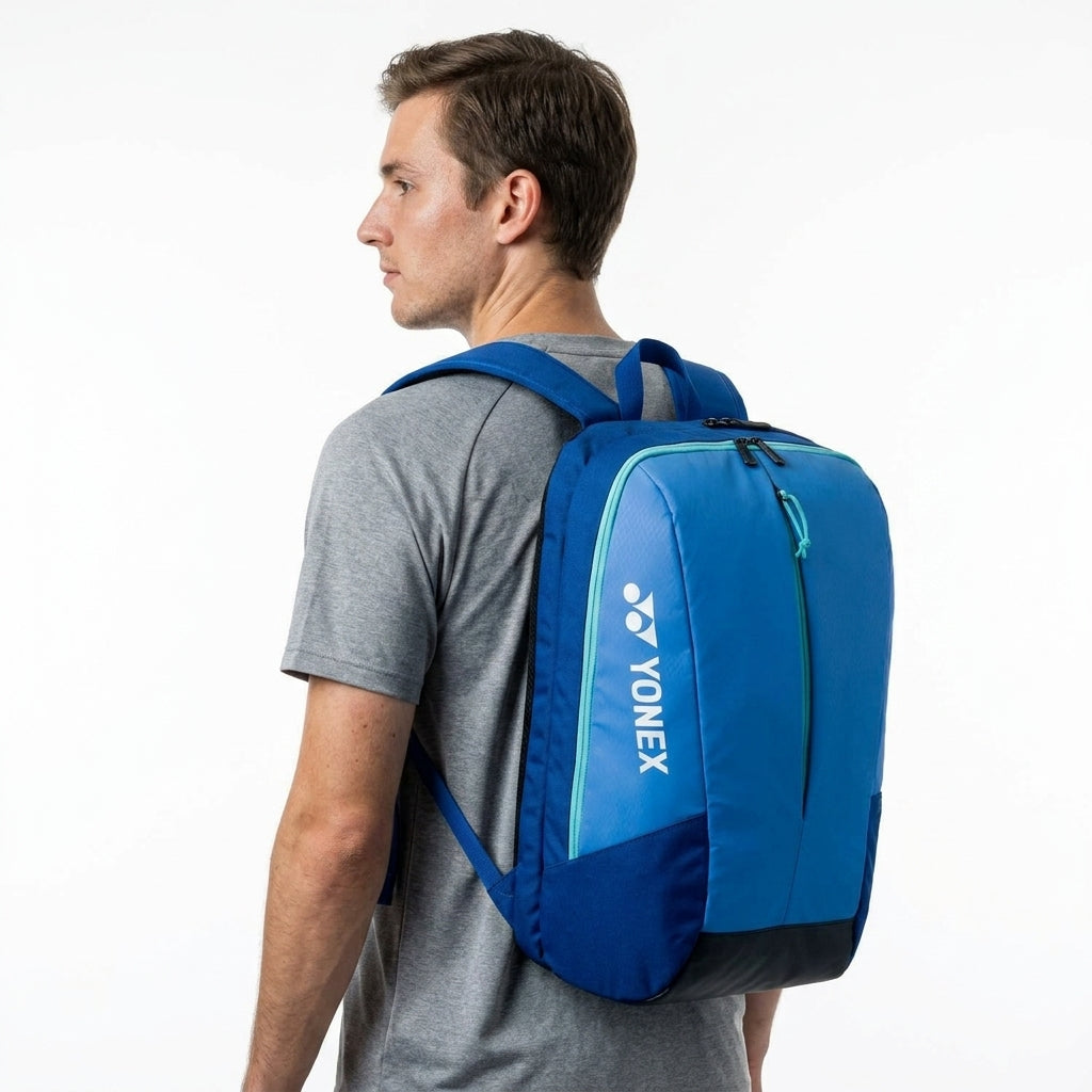 A man wearing a grey t-shirt and carrying a Yonex 42512EX Team Badminton Backpack in Blast Blue stands facing left against a plain white background.
