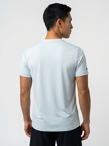 A man with short black hair stands with his back to the camera, wearing a Babolat Play Men's Crew Neck Badminton T-Shirt in Illusion Blue, highlighting stylish Babolat badminton apparel against a plain light background.