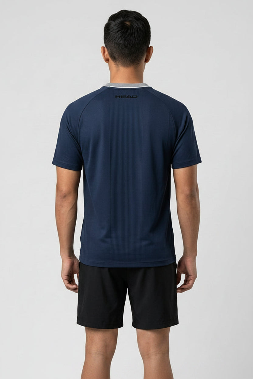 A man faces away from the camera, modeling a HEAD Performance Mens Badminton T-Shirt in navy and black shorts against a light gray background. The moisture-wicking shirt features "HEAD" printed below the neckline.