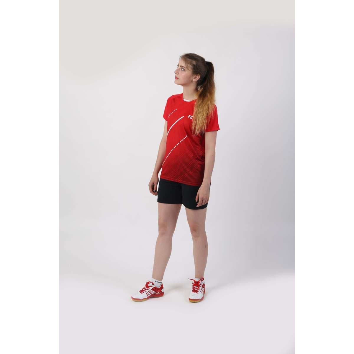 A person stands looking up, dressed in a vibrant FZ Forza Bali Red Girls/Women's Badminton T-Shirt, black shorts, and matching red sneakers. The comfortable ensemble contrasts against the plain white background.