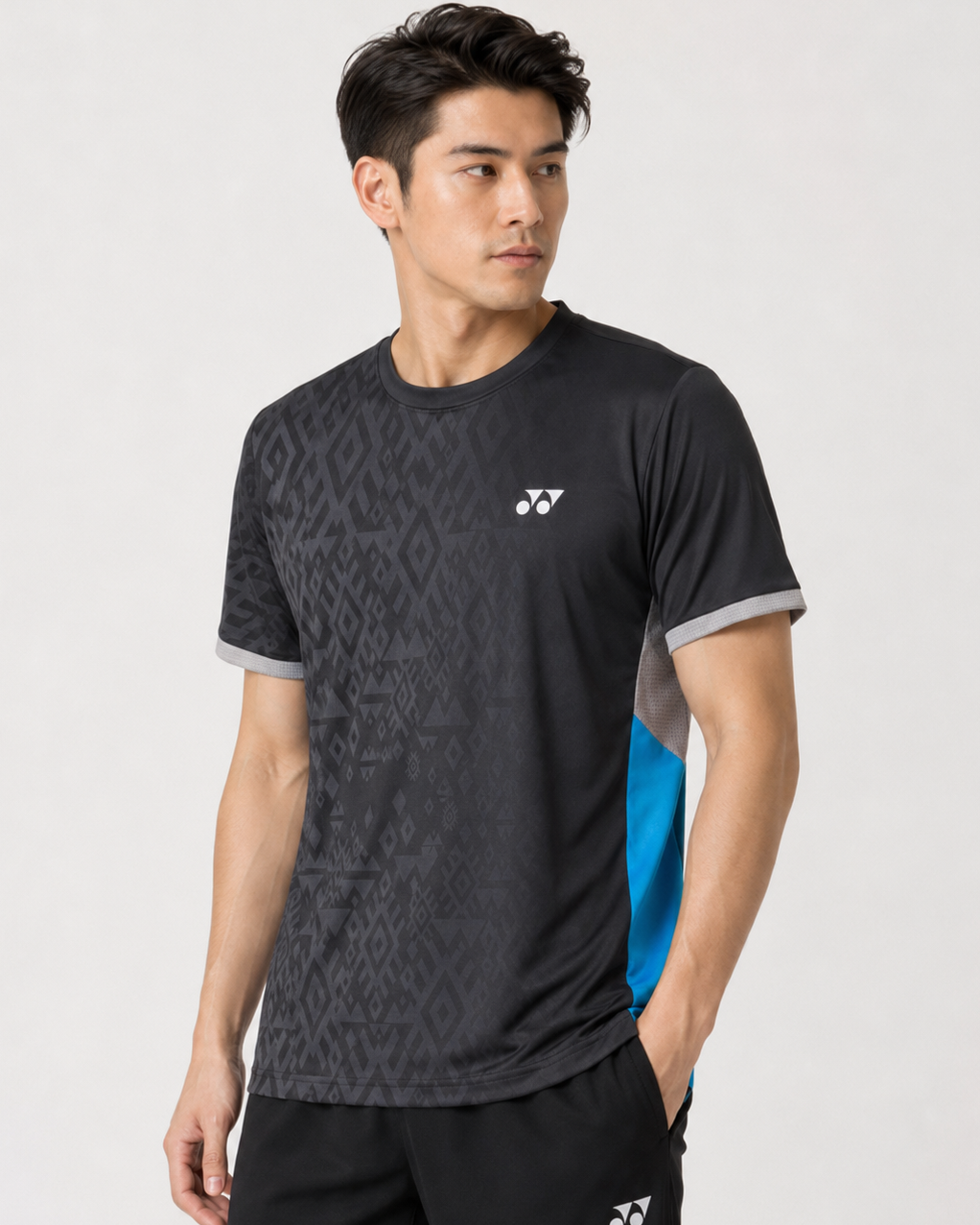 A man wearing the Yonex YTM11 Men's Badminton T-Shirt in black by Yonex, featuring gray trim and blue side panels in Air-Release fabric, stands with one hand in his pocket, looking left against a plain light background.