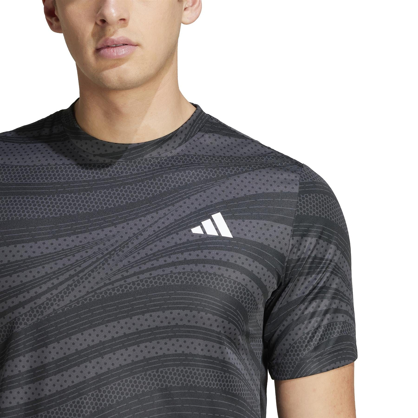 A person wears an ADIDAS Men's Club Graphic Badminton T-Shirt by adidas, made from 100% recycled materials. The black shirt features gray patterns and a white geometric logo on the chest, highlighting its stylish, eco-friendly design in the image.