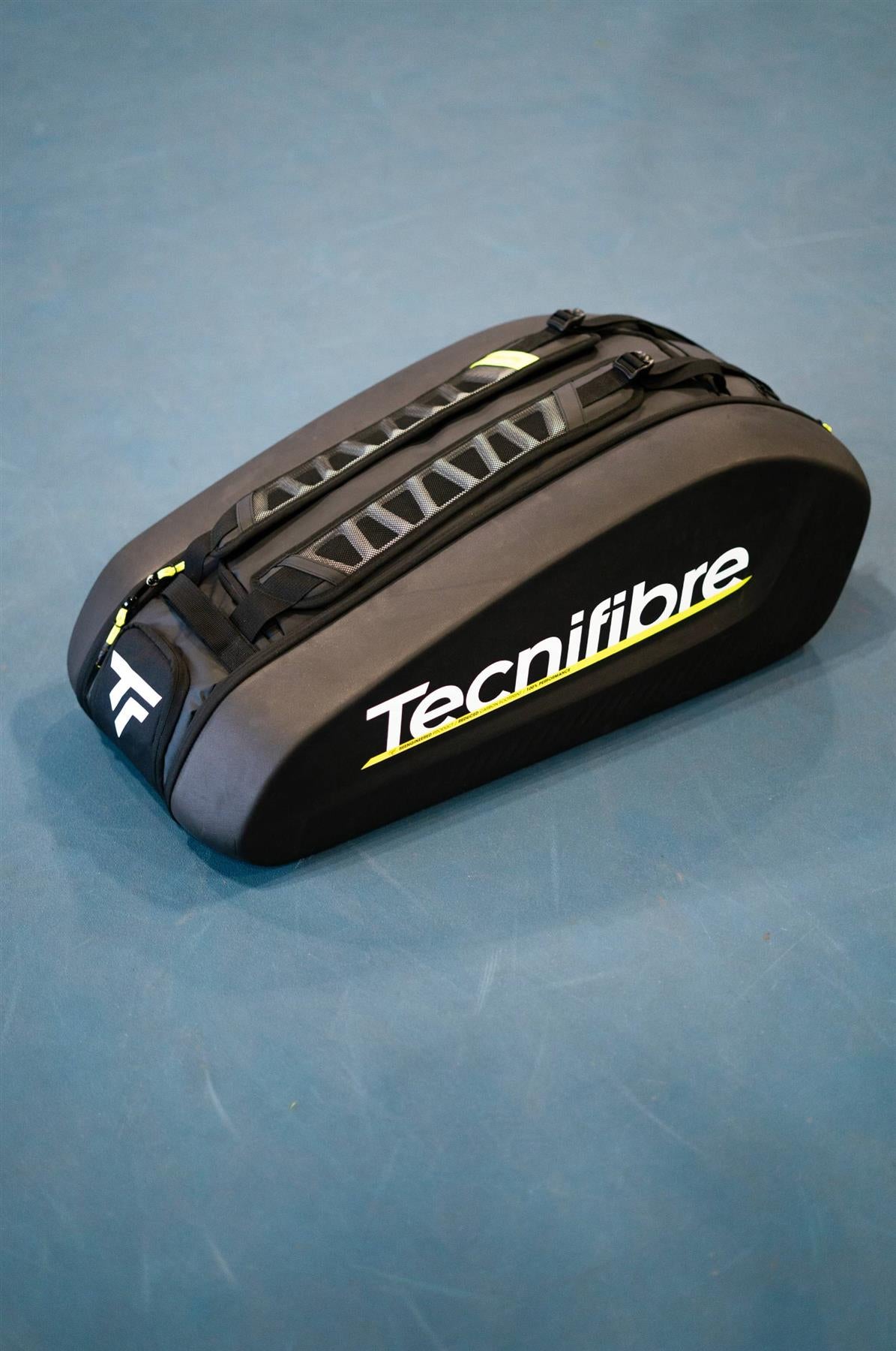 The Tecnifibre Reform L Badminton Racket Bag - Black features yellow and white accents, a large logo, top carrying straps, and spacious EVA compartments, shown here on a blue tennis court.