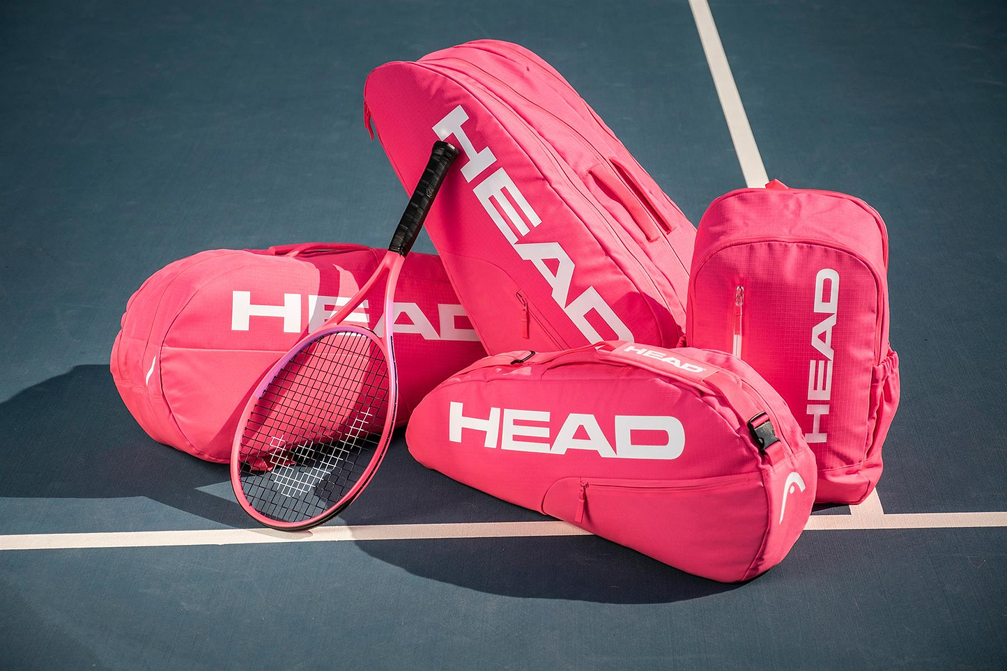 Four vibrant pink HEAD Base 3 Racket Badminton Bag S by HEAD and a matching racket are displayed on a blue tennis court, with the bags positioned on and around the white lines.