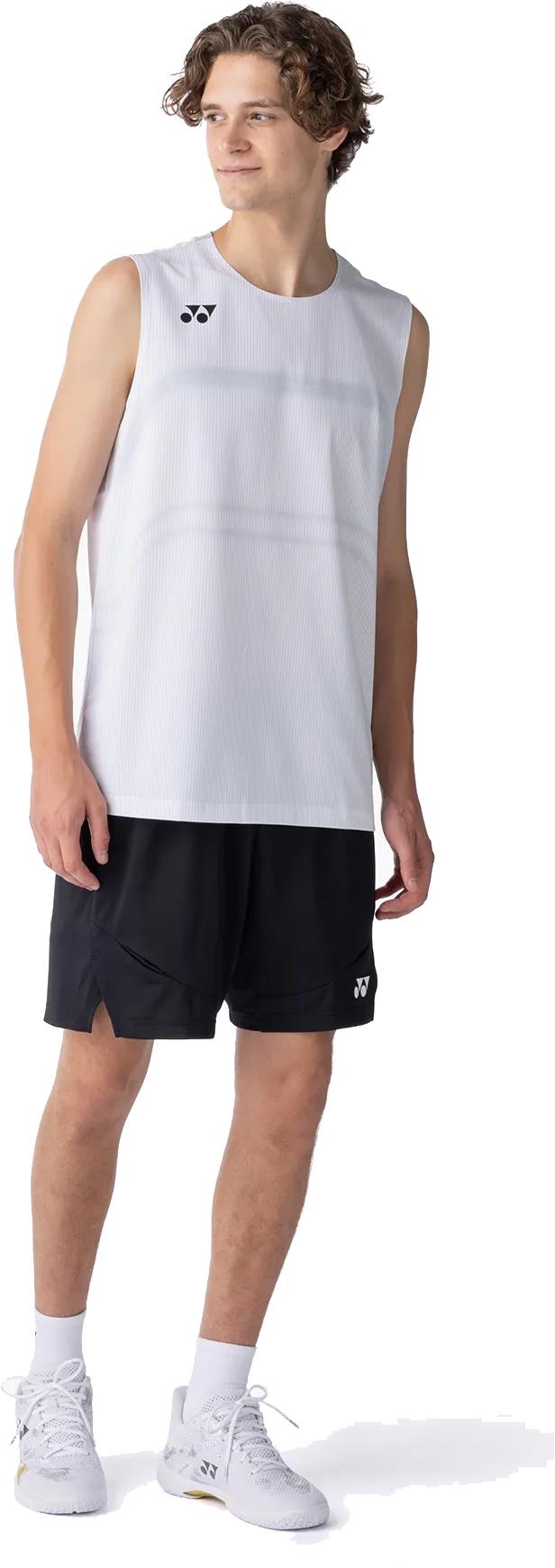 A young man in a sleeveless white athletic shirt, Yonex 15239YX Knit Men's Badminton Shorts in black, white socks, and white sneakers stands slightly turned to the side, looking off-camera against a white background.