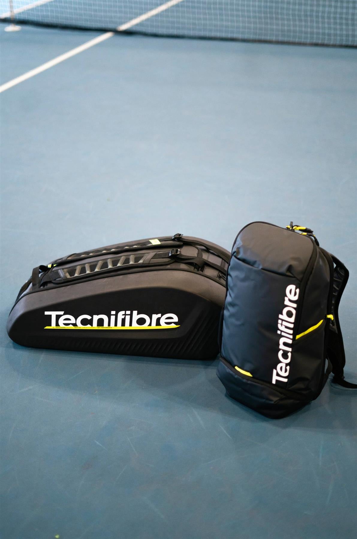 Two black Tecnifibre Reform L Badminton Racket Bags with white Tecnifibre logos and yellow accents rest on a blue tennis court near the net. These bags are spacious and made with durable EVA material.