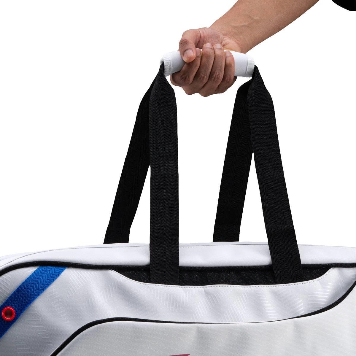 A hand holds the white handle and black straps of the Li-Ning Competition 6 Racket Badminton Bag (White/Black) by Li-Ning, shown against a plain white background.