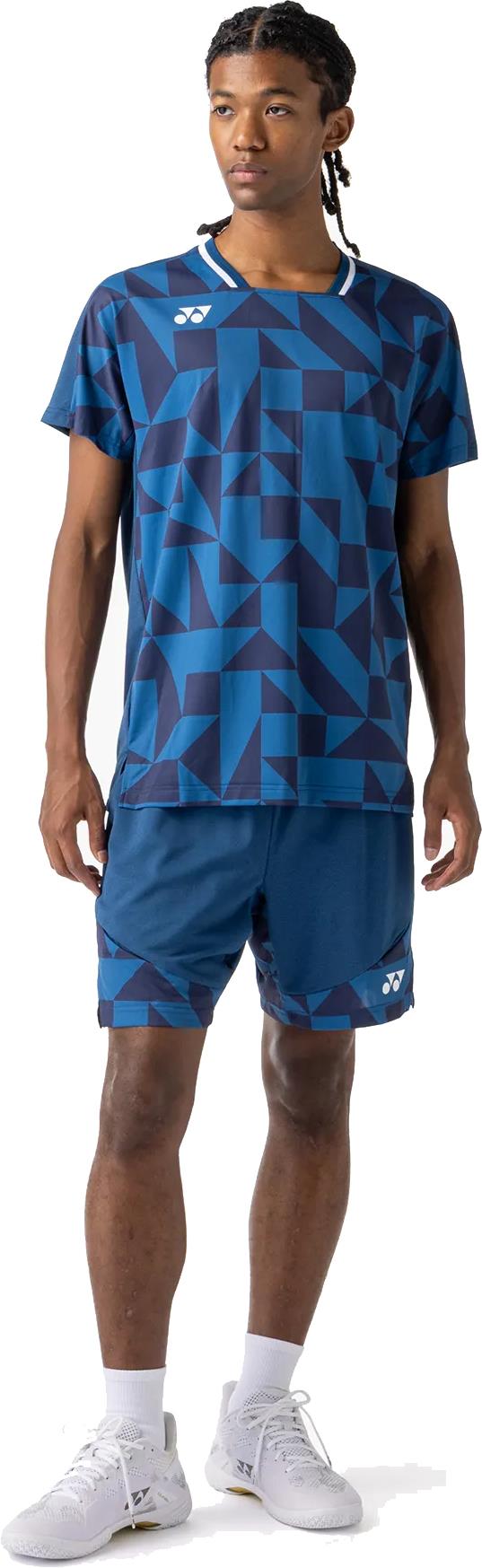 A man with braided hair, looking to the side, wears a blue geometric-patterned athletic shirt and Yonex 15240YX Knit Men's Badminton Shorts in Indigo Blue, paired with white socks and athletic shoes.