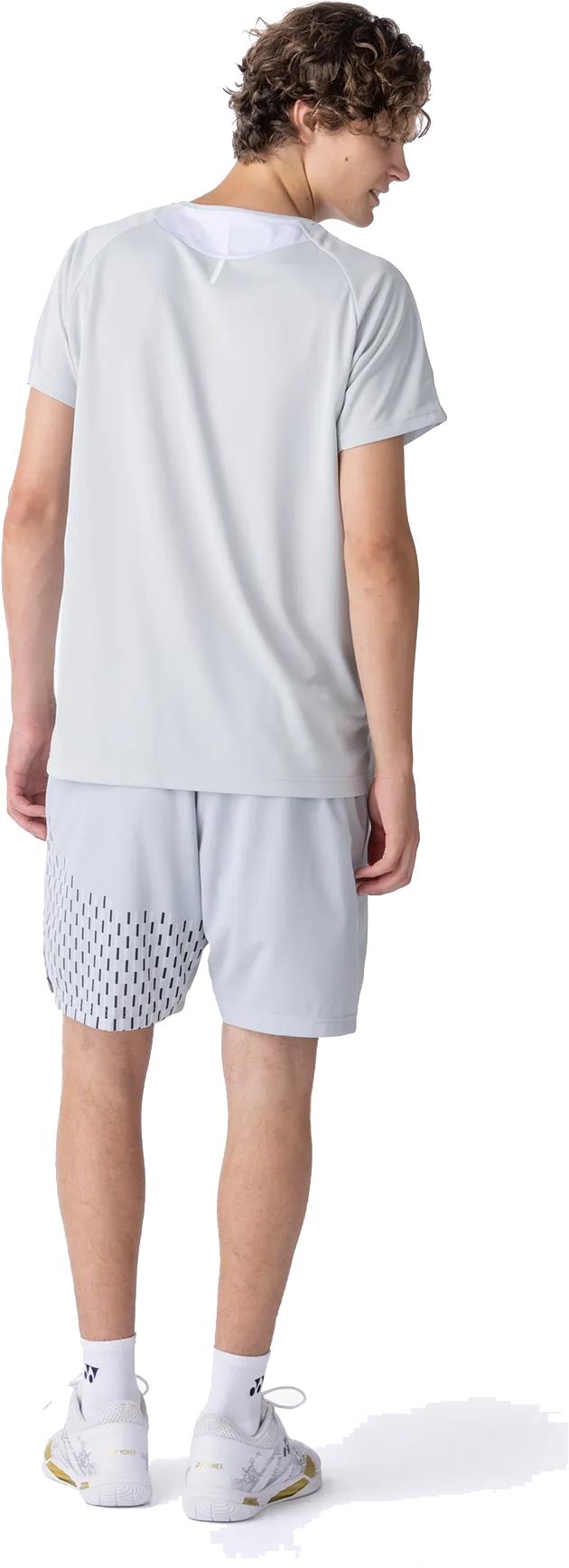 A person with curly hair faces away, wearing a light gray t-shirt and Yonex 15241EX Knit Men's Badminton Shorts in Silver Grey, paired with white socks and athletic shoes. The background is plain white.