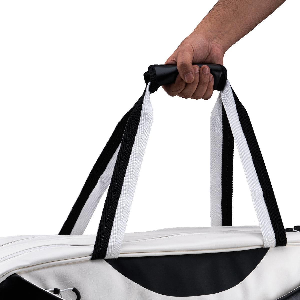 A person's hand holds the Li-Ning Competition 6 Racket Badminton Bag in white with black accents, a duffel-style bag by Li-Ning designed to carry up to 6 rackets, shown against a plain white background.