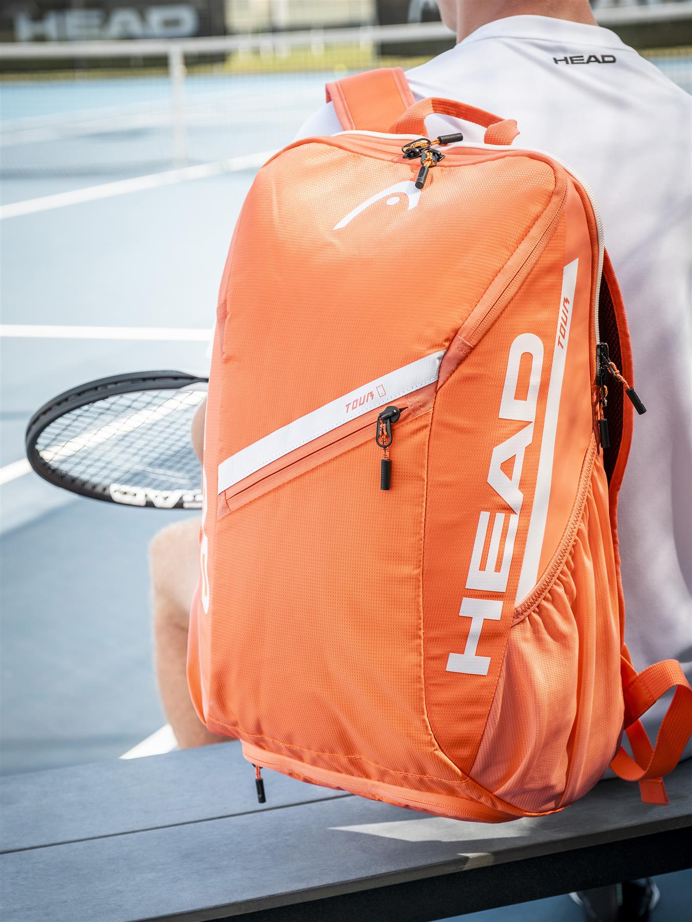 A HEAD Tour 25L Badminton Backpack in orange sits on a bench beside someone holding a racket, with a blurred tennis court and net in the background.