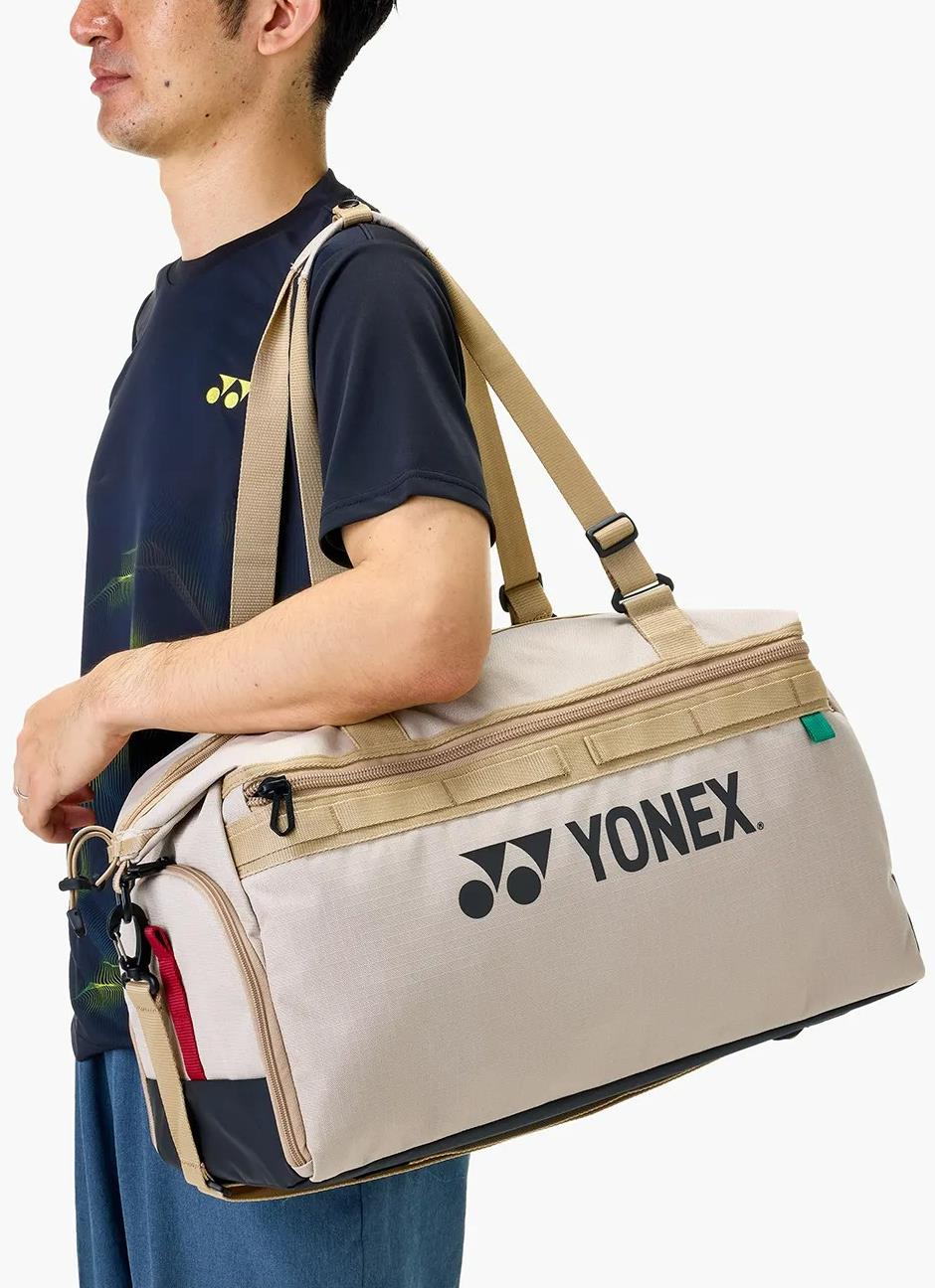 A person in a dark sports shirt carries the Yonex BA72611EX Gearlogic Badminton Boston Bag in beige, featuring tan straps and the Yonex logo. Made from recycled polyester, it's ideal for eco-conscious athletes.