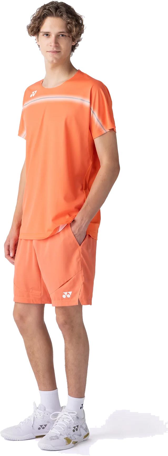 A young man faces forward, wearing the Yonex 10711YX Crew Neck Mens Badminton T-Shirt in Cyber Orange with matching shorts, white logo, socks, and athletic shoes. His left hand is in his pocket, highlighting this performance sportswear look.
