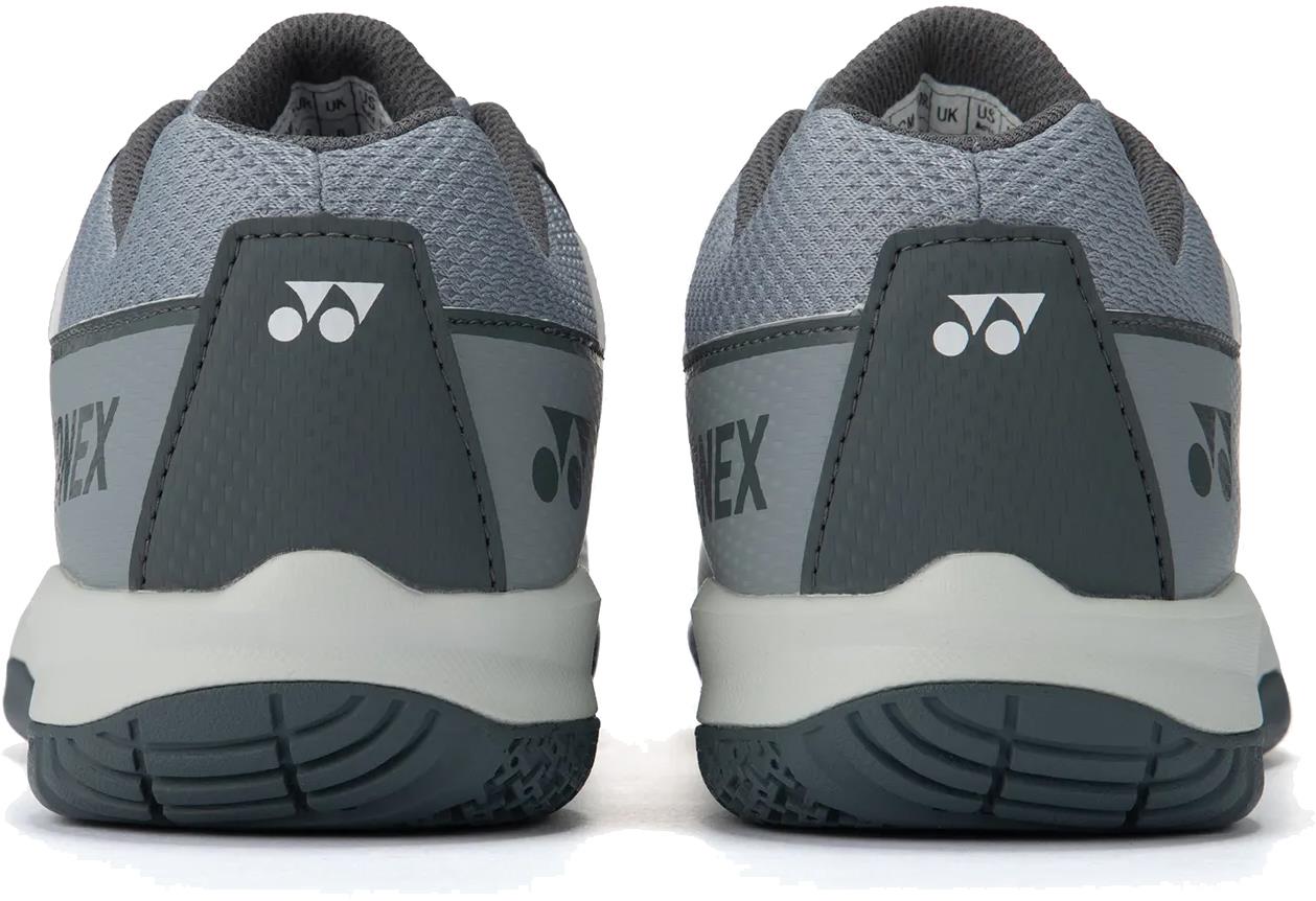 A close-up rear view of light grey Yonex Strider Flow unisex badminton shoes, featuring textured soles, Yonex branding, and a triangular logo on each heel.