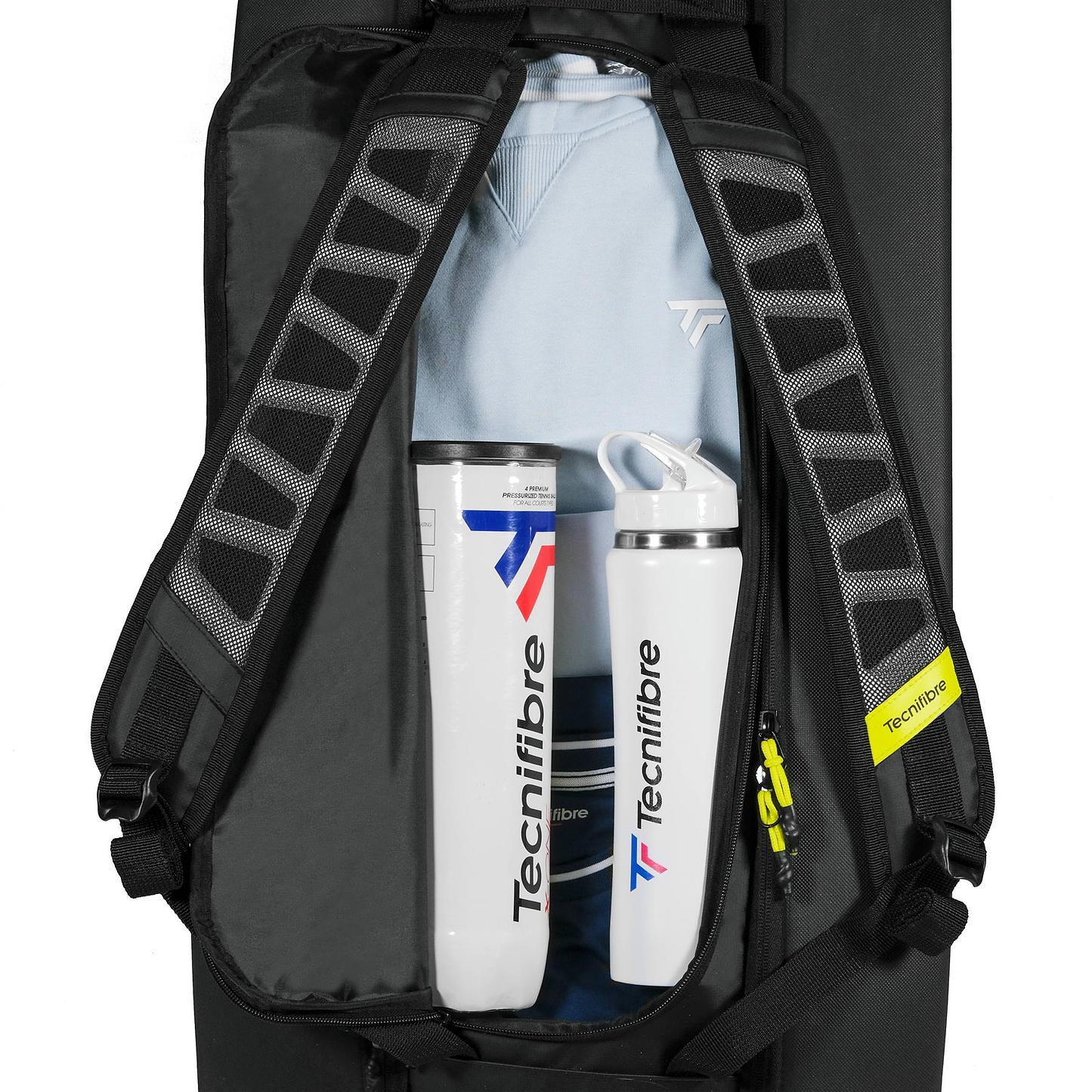 An open Tecnifibre Reform L Badminton Racket Bag in black shows two Tecnifibre water bottles, a folded light blue sweatshirt, and other gear inside. A yellow Tecnifibre tag hangs from one of the bag’s roomy straps.