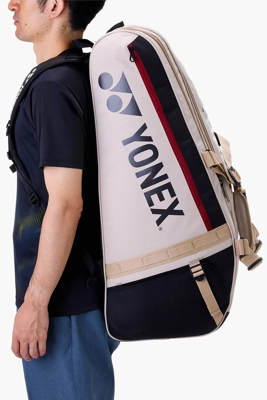 A person is seen from the side wearing a spacious Yonex BA72629EX Gearlogic 9 Racket Badminton Bag in beige and black with a red zipper, featuring the prominent Yonex logo and name on the back.