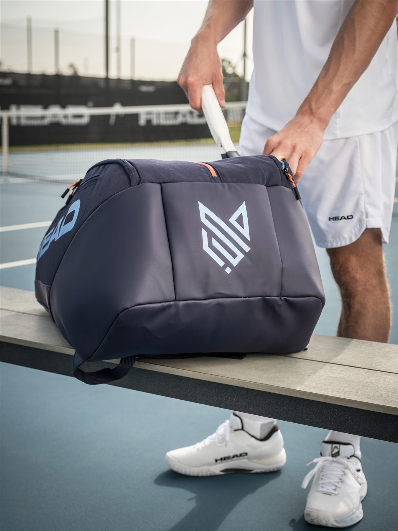 A tennis player in white sportswear places a racket into the large navy HEAD Pro 12 Racket Badminton Bag XL with CCT+ climate control by HEAD, resting on a bench with badminton courts visible in the background.