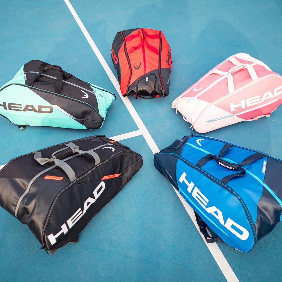 Five sports bags from the HEAD Tour Team 12R Monstercombi collection are neatly arranged on a blue tennis court. These colorful racket bags are available in mint green, red, pink, black, and blue shades and prominently feature "HEAD" in large white letters. They also incorporate CCT+ climate control technology to safeguard your gear.