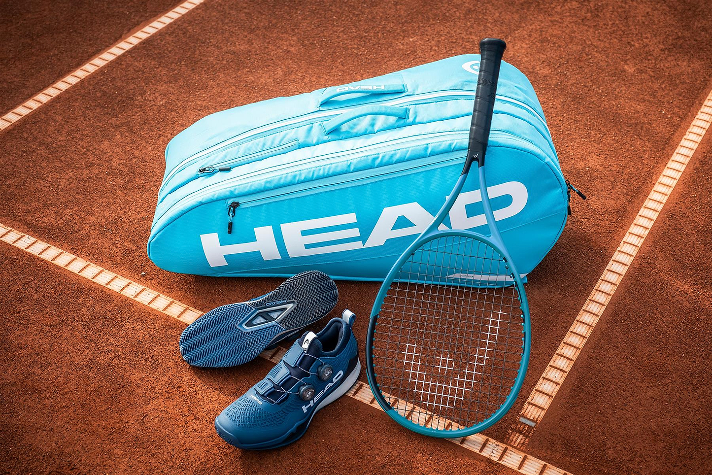 A HEAD Tour 9 Racket Badminton Bag L - Blue, a matching racket, and blue tennis shoes are placed on a clay court near the sideline.