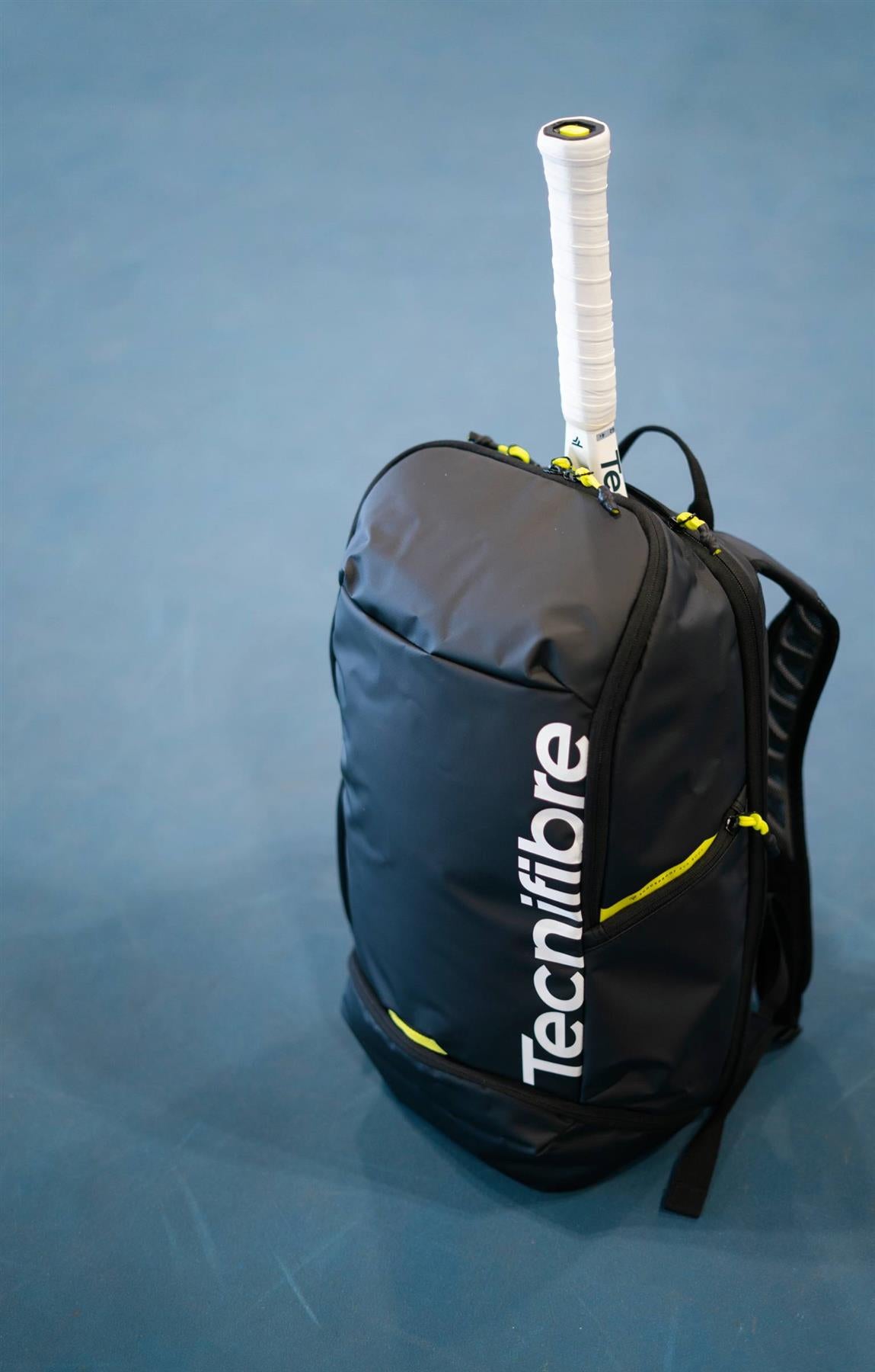 The Tecnifibre Reform Badminton Backpack - Black sits on a blue court, yellow accents highlighting its versatile design as it holds a tennis racket with a white grip sticking out from the top.