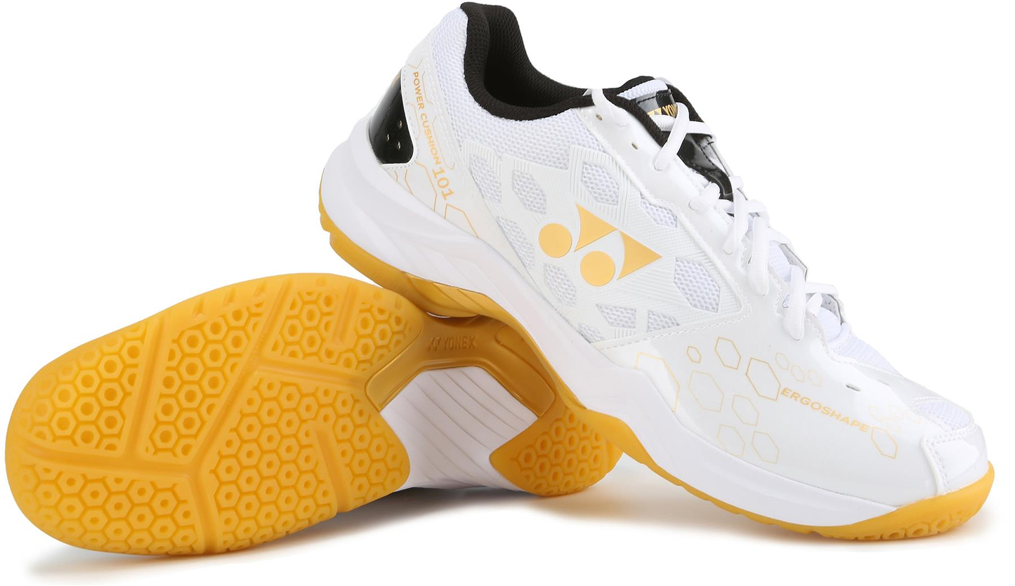 The Yonex Power Cushion SHB 101 Badminton Shoes in white and gold feature a honeycomb design for superior shock absorption, yellow-gold accents, visible tread pattern, and the Yonex logo on the side. One shoe stands upright; the other is on its side.