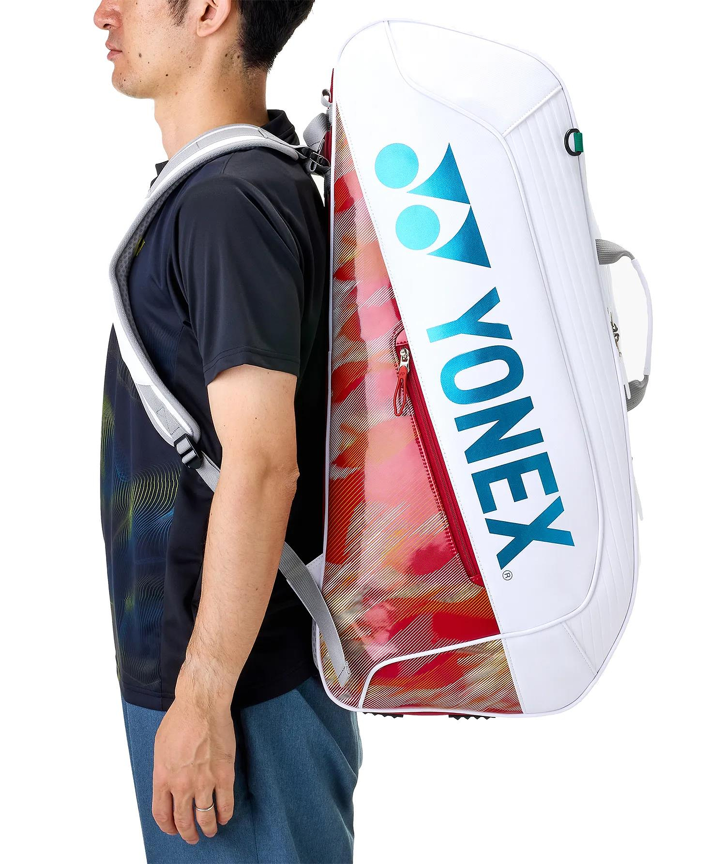 A man in a black shirt and blue shorts carries a Yonex 02626EX Expert 6 Racket Badminton Bag in white with red accents on his back, shown from the side.