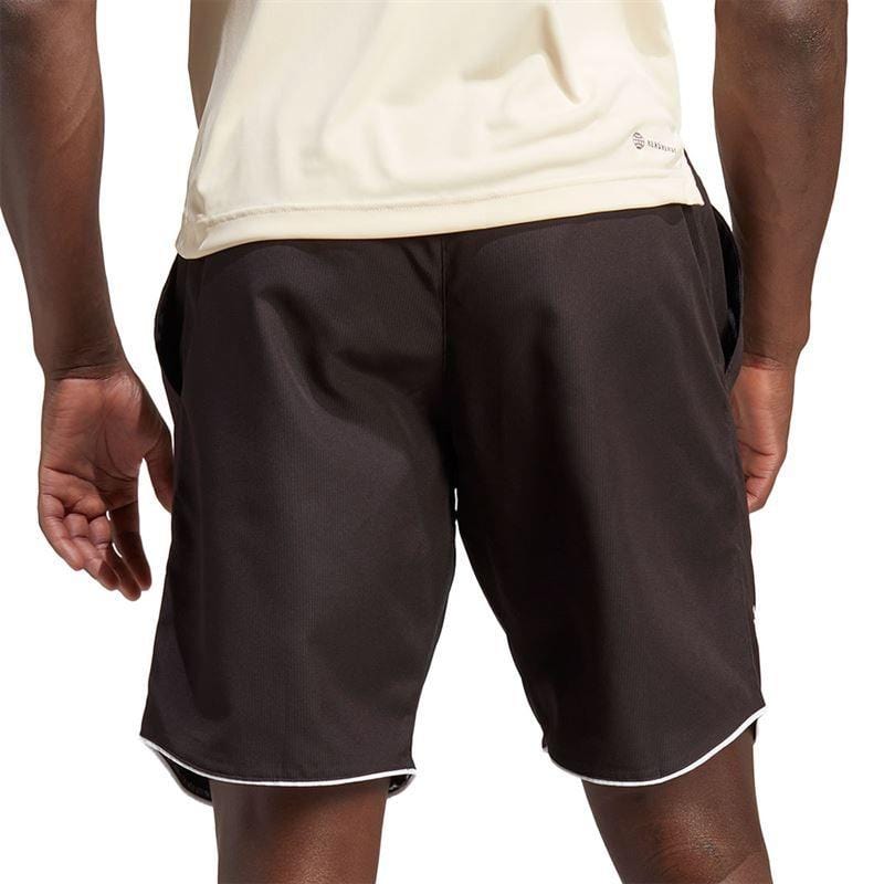 A person wearing a light-colored AEROREADY athletic shirt and the ADIDAS Men's Club 7" Badminton Shorts in black with white trim stands with their back to the camera, hands relaxed by their sides. The plain white background accentuates the sleek design of their attire.