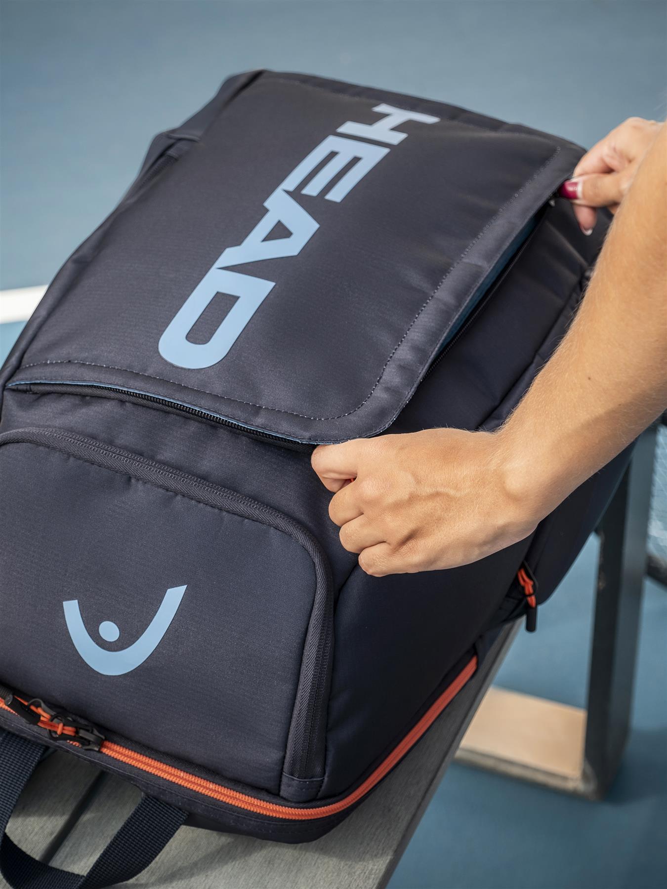 A person opens the top racket compartment of a navy HEAD Pro 28L Badminton Backpack with light blue lettering and orange zipper accents, placed on a gray bench with a tennis court in the background.