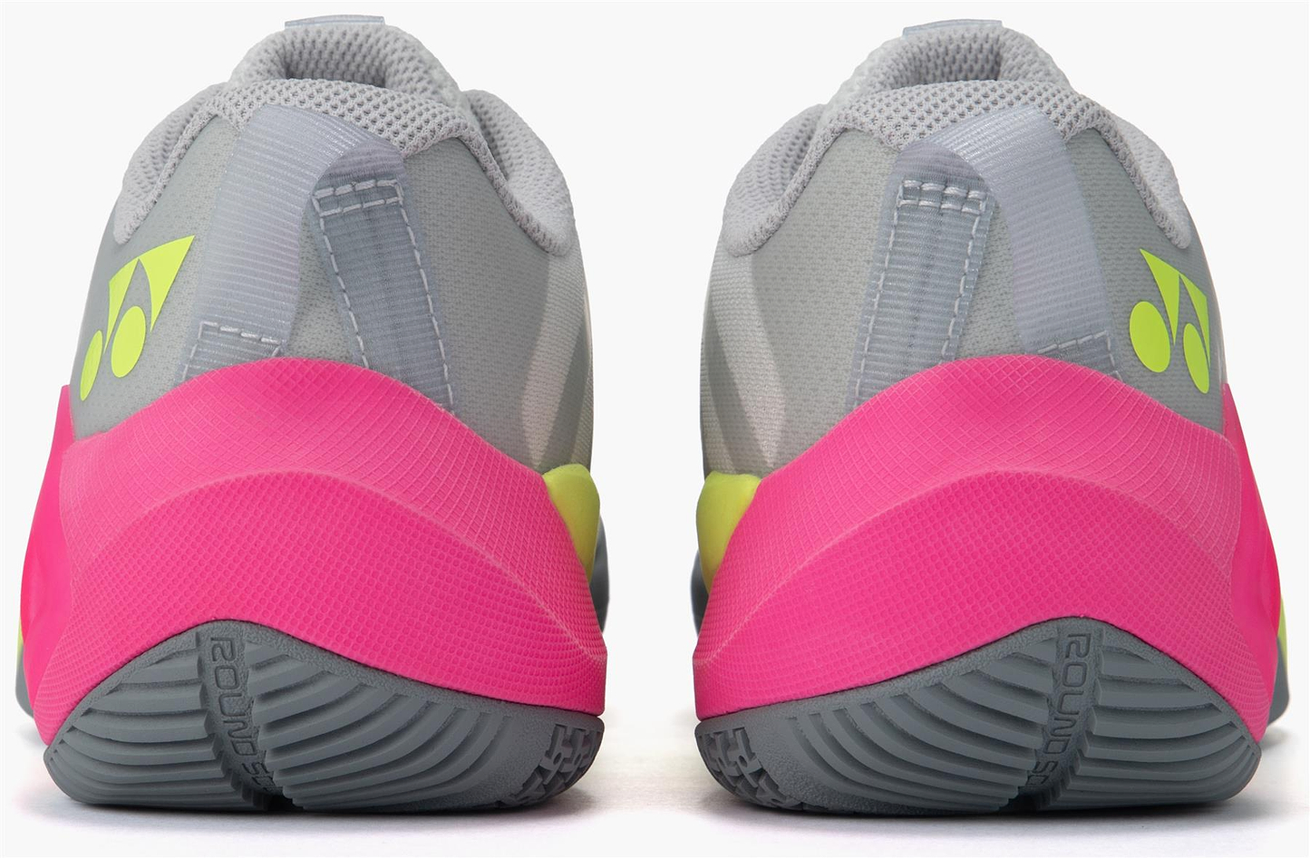 The Yonex Subaxia GT Women's Badminton Shoes in light grey feature bright pink heel accents, yellow Yonex logos, textured dark grey soles, and advanced GRPHT THRTTL technology for enhanced performance.