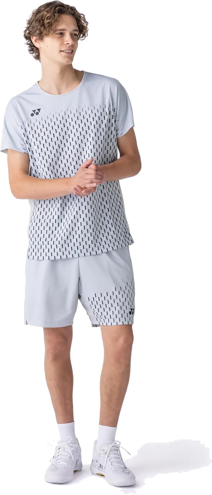 A young man wears a light gray athletic set with black dash patterns, featuring a Yonex 15241EX Knit Mens Badminton Shorts in Silver Grey and white sneakers. He looks to the side, hands clasped.